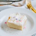 A white frosted with sprinkles yummy sugar cookie bar on a white plate with gold forks in the background.