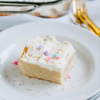 A white frosted with sprinkles yummy sugar cookie bar on a white plate with gold forks in the background.