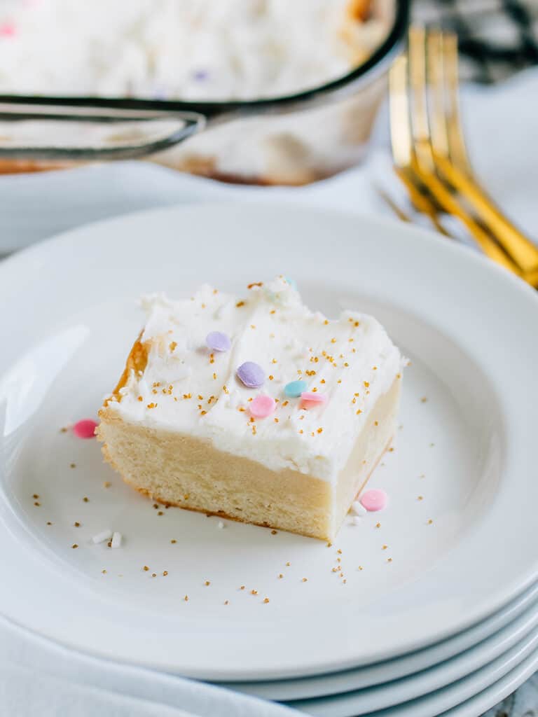 A white frosted with sprinkles yummy sugar cookie bar on a white plate with gold forks in the background.
