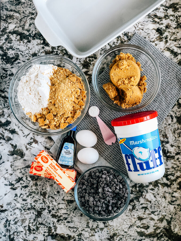 Ingredients for 9x13 inch S'mores Cookie Bars: Crushed graham crackers, flour, baking powder, eggs, vanilla, butter, brown sugar, marshmallow creme, and chocolate chips.
