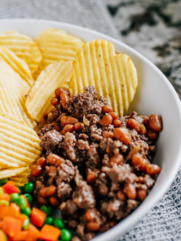 A plate of hamburger and baked beans with chips and steamed vegetables on the side.