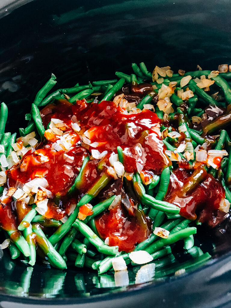 The BBQ sauce, bacon and onions on top of the layer of green beans in a crockpot.
