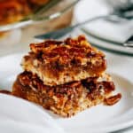 Two pecan pie bars stacked on top of each other showing the crisp crumble crust, sweet filling, an candy pecans of these amazing pecan pie bars.