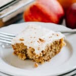 A apple blondie with maple cream cheese frosting on a serving plate with apples in the background.