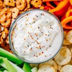 Super close up of Everything Bagel Dip in a glass bowl on a large wooden cutting board surrounded by pretzel crisps, bell peppers, pita crackers, and celery.