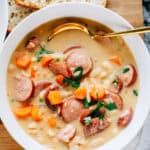 Overhead view: A bowl of savory kielbasa, soft cooked carrots, and creamy Great Northern beans simmered to perfection in a cream sauce with minced garlic and onions. This is the best bean soup! Our famous homemade peasant bread on the side.