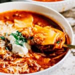Side view with a spoonful of soup being lifted out of the white soup bowl. A bowl of warm and comforting lasagna soup with beef in a beef and marinara broth topped with a dollop of ricotta cheese, shredded cheddar cheese and some parsley.