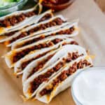 A large cutting board lined with parchment paper and bowls of chopped lettuce, fresh salsa, sour cream surrounding oven-hot ground beef street tacos.