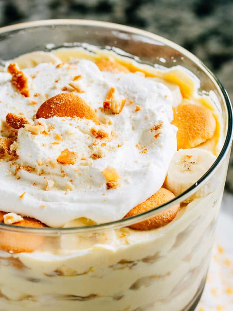 Close up: Layers of vanilla pudding, crushed Nilla wafers, and sliced bananas in a trifle bowl with whipped cream topping. This Banana Pudding dessert is fun and delicious!