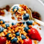 A spoonful of homemade cinnamon sugar granola, fresh blueberries, and yogurt raised above a bowl on the same for a colorful and flavorful snack or breakfast.