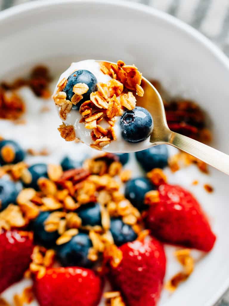 A spoonful of homemade cinnamon sugar granola, fresh blueberries, and yogurt raised above a bowl on the same for a colorful and flavorful snack or breakfast.