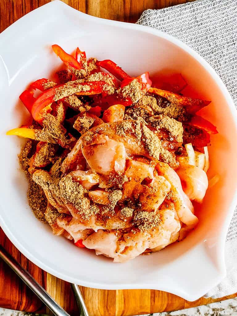 Raw sliced chicken, peppers, and onions with oil and fajita seasoning on top in a large mixing bowl.