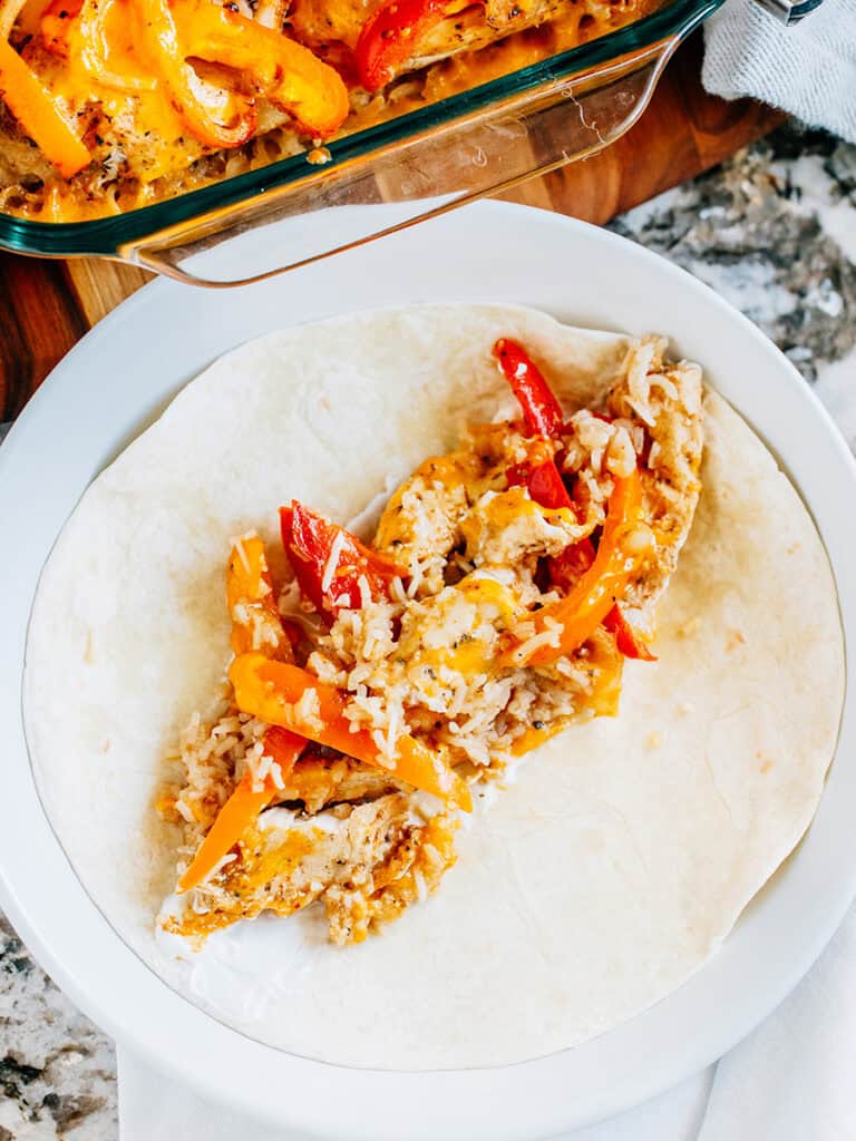 A flour tortilla with sour cream down the middle and this chicken fajita bake for a simple chicken fajitas taco. Baking dish of chicken fajitas with cheese in backgrounds.