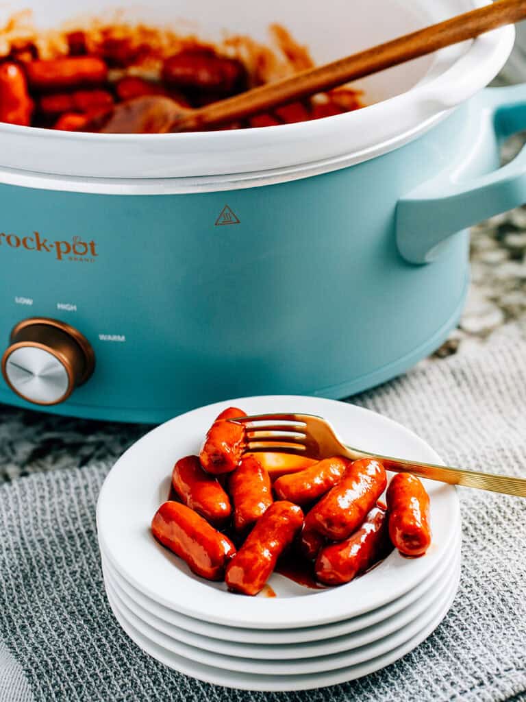 A plate of li'l smokies coated in a savory bbq sauce sitting in front of a slow cooker ready to be eaten.