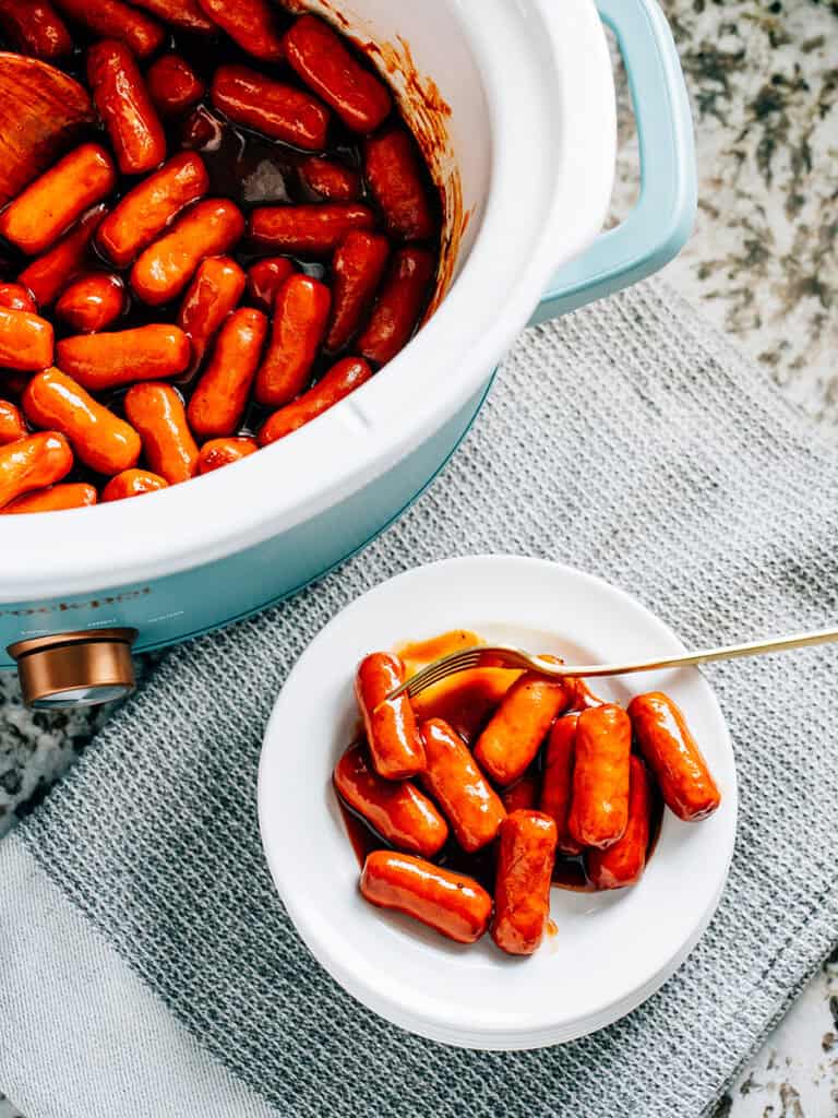 Overview shot: A plate of li'l smokies coated in a savory bbq sauce sitting in front of a slow cooker ready to be eaten.