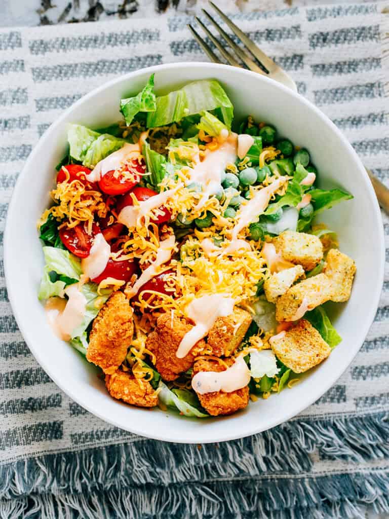 A delicious buffalo chicken salad with crisp romaine, warm chicken chunks, cherry tomatoes, frozen peas, cheddar cheese, croutons, and a drizzle of buffalo dressing.