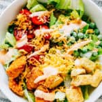A delicious buffalo chicken salad with crisp romaine, warm chicken chunks, cherry tomatoes, frozen peas, cheddar cheese, croutons, and a drizzle of buffalo dressing plated and ready to serve