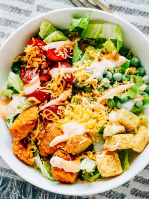A delicious buffalo chicken salad with crisp romaine, warm chicken chunks, cherry tomatoes, frozen peas, cheddar cheese, croutons, and a drizzle of buffalo dressing plated and ready to serve