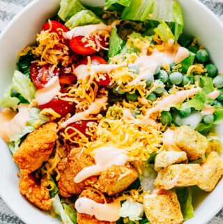 A delicious buffalo chicken salad with crisp romaine, warm chicken chunks, cherry tomatoes, frozen peas, cheddar cheese, croutons, and a drizzle of buffalo dressing plated and ready to serve