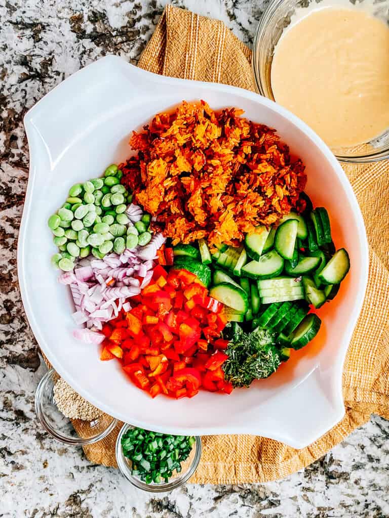 A large bowl with shelled edamame, cucumbers, red peppers, red onion, parsley and crispy rice ready to be assembled into a salad. A bowl of homemade spicy mayo dressing on the side. The toppings on the side as well in small glass containers.