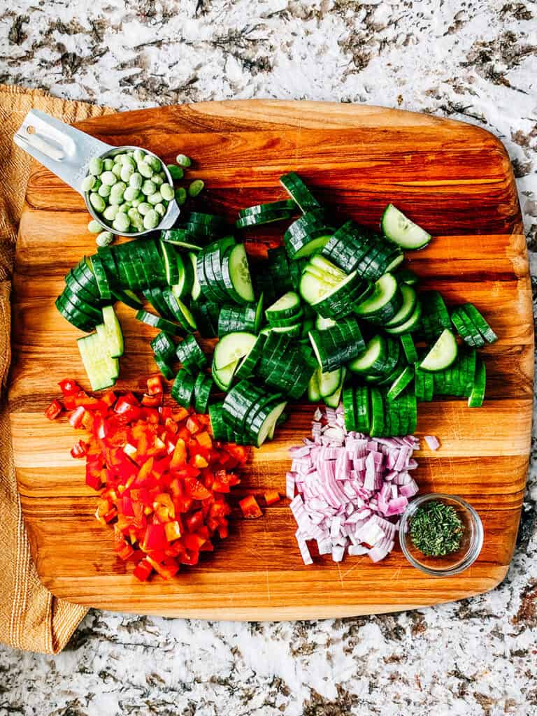 A cutting board showing the salad ingredients: Frozen shelled edamame, sliced cucumbers, diced red peppers, red onions, and parsely.
