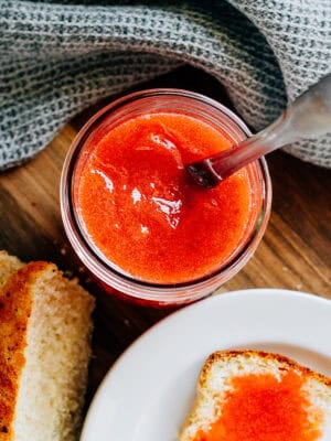 A jar of delicious homemade strawberry jam with a knife in it and some of it spread on a piece of bread on the side.