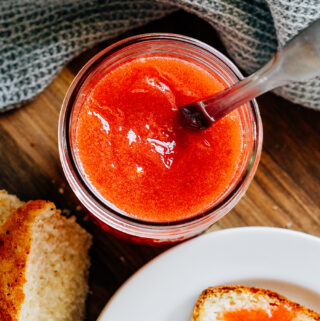 A jar of delicious homemade strawberry jam with a knife in it and some of it spread on a piece of bread on the side.
