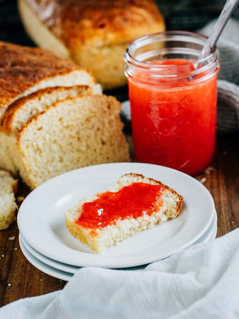 A piece of cottage cheese bread with sweet strawberry jam spread on it. A jar of jam in the background with a loaf of homemade bread.