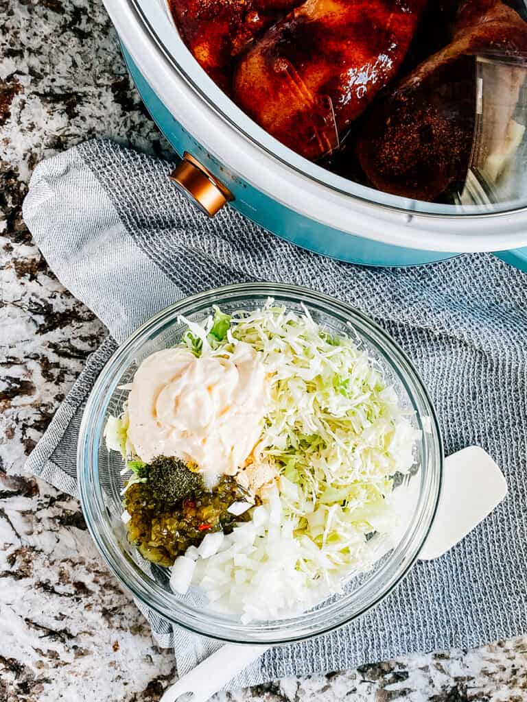 Next to the slow cooker is a glass bowl with the pickle slaw ingredients: Shredded cabbage, mayo, diced sweet onion, pickle relish, garlic powder, salt, and dill.