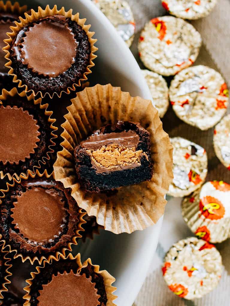 A peanut butter cup brownie bite cute in half to show off the chocolate brownie and peanut butter inside surrounded but more brownie bites on one side and Reeses mini peanut butter cups on the other side.