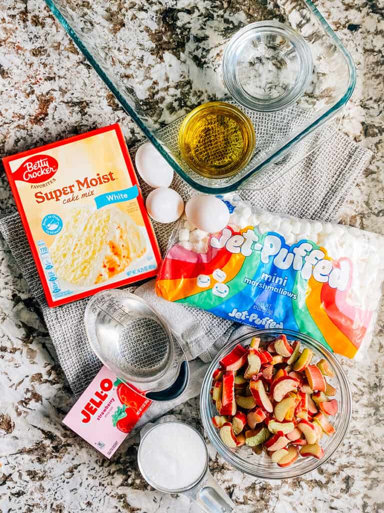 The ingredients to this strawberry jello rhubarb cake: White cake mix, eggs, oil, water, mini marshmallows, water for Jell-O, Strawberry Jell-O, sugar, and chopped rhubarb.