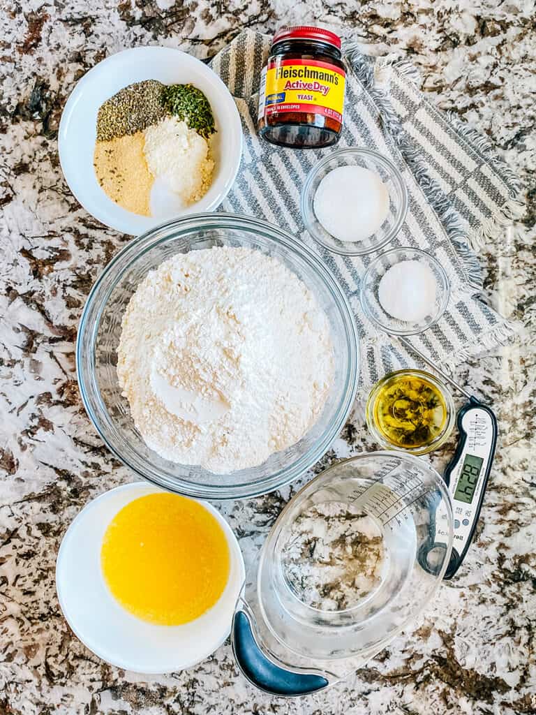 Ingredients for homemade breadsticks with a garlic seasoning: four, sugar, salt, yeast, warm water, oil, butter, grated parmesan, dried oregano, basil, parsley, garlic powder, and salt.