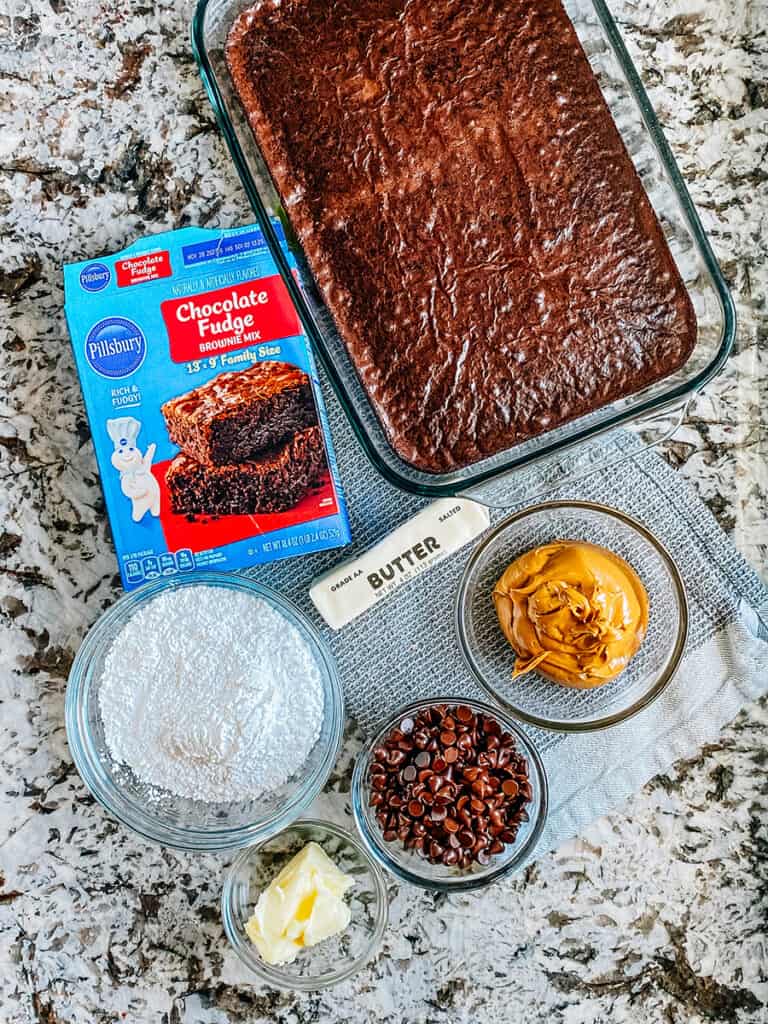 The ingredients to buckeye brownies using a box brownie mix. Baked box brownies, butter, peanut butter, powdered sugar, and semi-sweet chocolate chips.