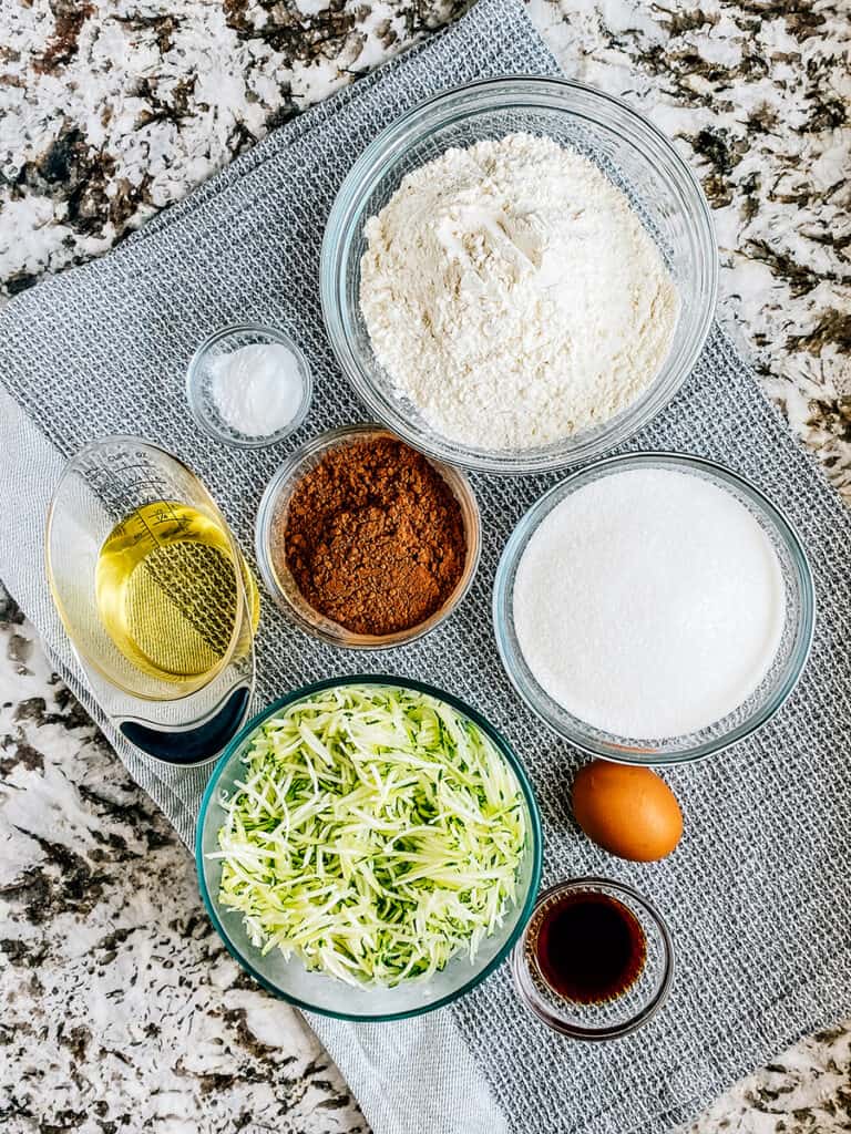 The ingredients to zucchini brownies: flour, sugar, baking soda, unsweetened cocoa powder, egg, oil, vanilla extract, and shredded zucchini.